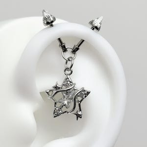 May include: A silver-toned industrial barbell piercing featuring a dangling star-shaped charm. The charm is decorated with small clear crystals and a central star cutout, suspended from the barbell by a small metal ring and two jump rings.