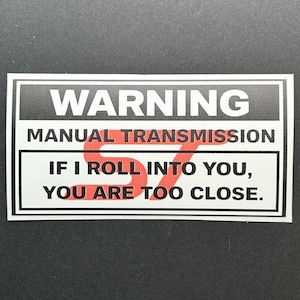 May include: A rectangular sticker with a black border and white background. The text reads "WARNING MANUAL TRANSMISSION IF I ROLL INTO YOU, YOU ARE TOO CLOSE." The word "ST" is in red, stylized font.