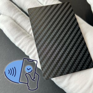 May include: A rectangular, carbon fiber card with a woven pattern. The card is dark gray and black. A blue graphic of a smartphone with a checkmark and a contactless payment symbol is in the lower left corner.