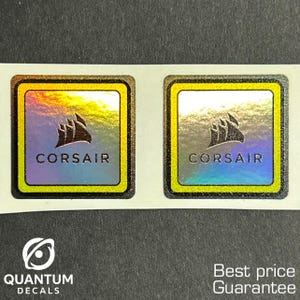 May include: Two square Corsair decals with a black logo and the word "CORSAIR" in white text. The decals have a holographic background and a gold border. The bottom of the image has the text "QUANTUM DECALS".