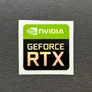 May include: A square sticker with the NVIDIA logo in green and white at the top. Below, the text "GEFORCE RTX" is printed in gold on a black background. The sticker is on a gray surface.