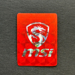 May include: A rectangular red sticker with a holographic logo. The logo features a dragon inside a shield, with the letters "MSI" below. The sticker has a textured, reflective surface.