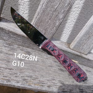 Peut inclure: Un couteau en acier inoxydable avec une lame incurvée et un manche avec un motif rouge et noir. Le texte "14C28N G10" est visible sur la surface en bois. Le couteau est conçu pour couper.
