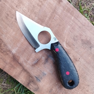 May include: A handmade knife with a polished steel blade and a black handle with two red dot accents. The blade has a circular cutout near the handle. The knife is resting on a wooden surface.