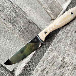 May include: A fixed-blade knife with a polished, reflective steel blade featuring a small logo near the bolster. The handle is made of a light beige, layered material with a textured grain and is secured by two dark circular rivets on the side.
