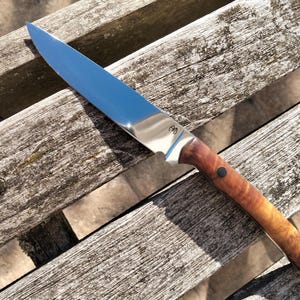 Può includere: Un coltello a lama fissa con una lama in acciaio lucida e appuntita e un manico in legno liscio color marrone chiaro. Il manico presenta due rivetti circolari scuri che fissano il legno al codolo in metallo. Un piccolo logo inciso è visibile sulla lama.