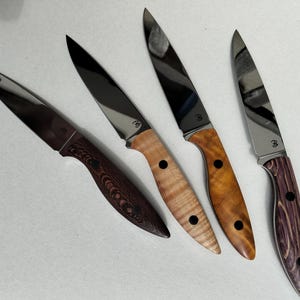 Può includere: Quattro coltelli con lame lucide e manici in legno di diverse tonalità di marrone e viola. Ogni coltello ha un piccolo punto scuro sul manico e una ghiera argentata. Le lame sono inclinate verso l'alto a destra.