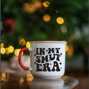 May include: White ceramic mug with a red rim and handle. The mug features the phrase "IN MY SMUT ERA" in black, stylized lettering. The mug is set against a blurred background with bokeh lights.