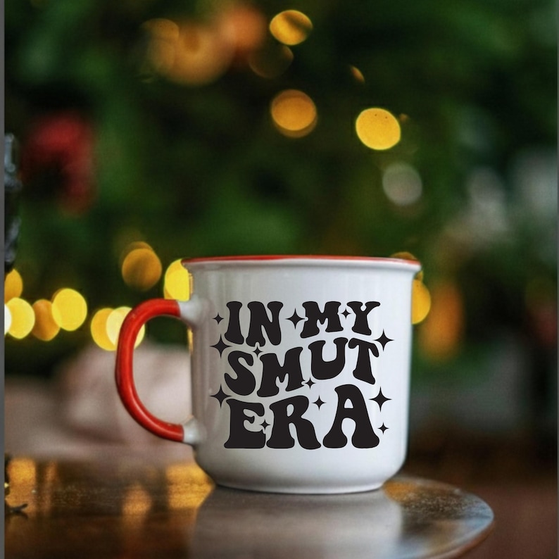 May include: White ceramic mug with a red rim and handle. The mug features the phrase "IN MY SMUT ERA" in black, stylized lettering. The mug is set against a blurred background with bokeh lights.