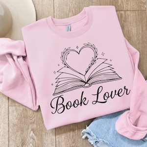 May include: A light pink sweatshirt with the words "Book Lover" in a cursive font. The graphic features an open book with a heart-shaped wreath above it. The design is in black and includes small star accents.