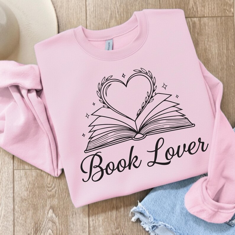 May include: A light pink sweatshirt with the words "Book Lover" in a cursive font. The graphic features an open book with a heart-shaped wreath above it. The design is in black and includes small star accents.