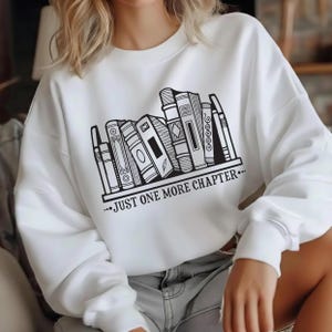May include: White sweatshirt featuring a black and white graphic of a bookshelf with various book designs. The text "...JUST ONE MORE CHAPTER..." is printed below the bookshelf. The sweatshirt is a casual style.