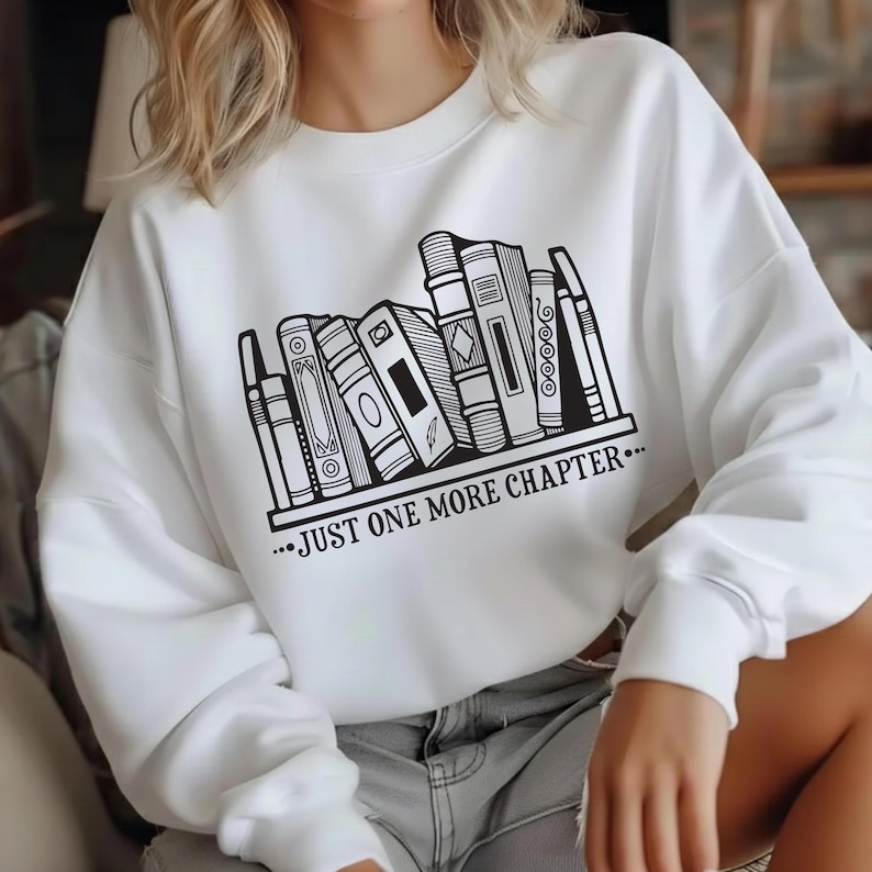 May include: White sweatshirt featuring a black and white graphic of a bookshelf with various book designs. The text "...JUST ONE MORE CHAPTER..." is printed below the bookshelf. The sweatshirt is a casual style.