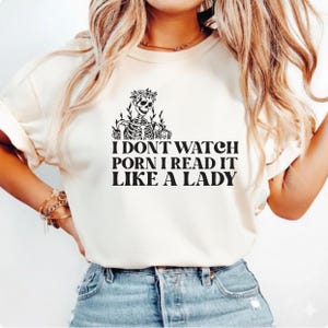 May include: Cream-colored t-shirt featuring a skeleton wearing a floral crown and holding a mug. The shirt has the text "I DON'T WATCH PORN I READ IT LIKE A LADY" in black, bold letters.