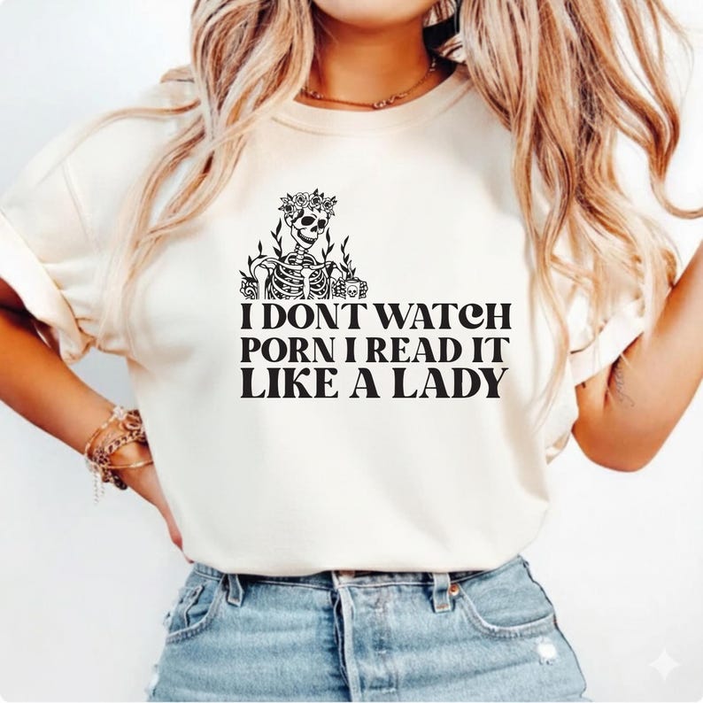 May include: Cream-colored t-shirt featuring a skeleton wearing a floral crown and holding a mug. The shirt has the text "I DON'T WATCH PORN I READ IT LIKE A LADY" in black, bold letters.