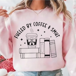 May include: A light pink sweatshirt with the phrase "FUELED BY COFFEE & SMUT" in black lettering. The graphic includes a coffee cup with a bow, steam, and books.