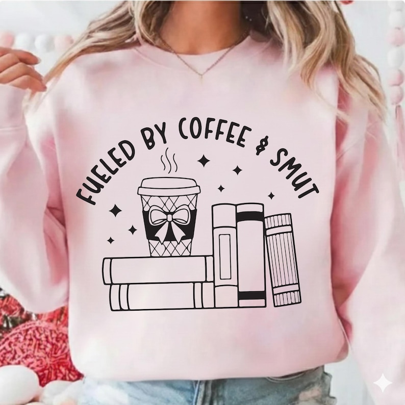 May include: A light pink sweatshirt with the phrase "FUELED BY COFFEE & SMUT" in black lettering. The graphic includes a coffee cup with a bow, steam, and books.