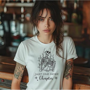 May include: White t-shirt featuring a black skeleton reading books, with the text "JUST ONE MORE CHAPTER" and star accents. The shirt has short sleeves and a crew neck.