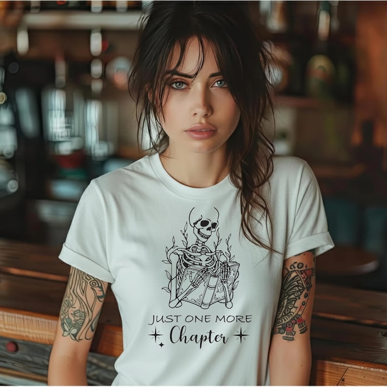 May include: White t-shirt featuring a black skeleton reading books, with the text "JUST ONE MORE CHAPTER" and star accents. The shirt has short sleeves and a crew neck.