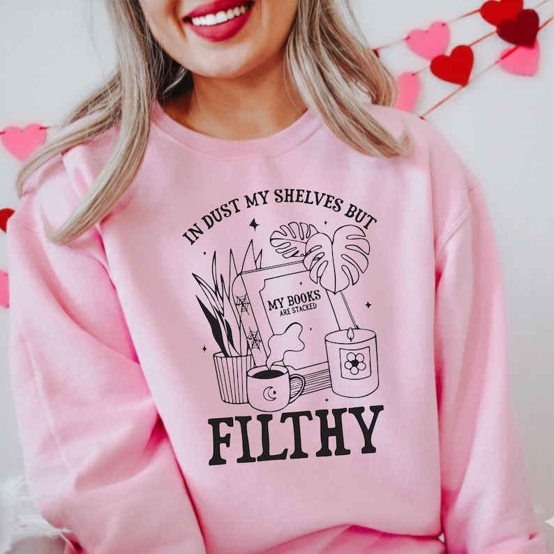 May include: Pink sweatshirt with a black graphic design. The design includes the text "IN DUST MY SHELVES BUT MY BOOKS ARE STACKED FILTHY" along with illustrations of plants, a book, a candle, and a coffee cup. The sweatshirt is a solid pink color.