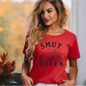 May include: A red t-shirt with the words "SMUT QUEEN" in black, above an open book illustration. The shirt is short-sleeved and has a crew neck. The design includes stars around the book.
