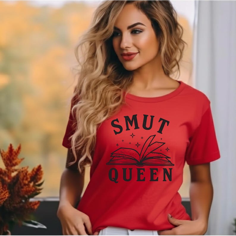 May include: A red t-shirt with the words "SMUT QUEEN" in black, above an open book illustration. The shirt is short-sleeved and has a crew neck. The design includes stars around the book.