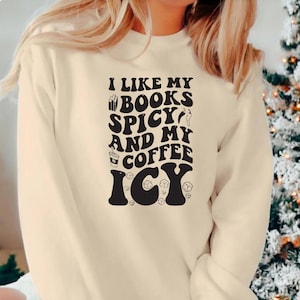 May include: Cream-colored sweatshirt with the text "I LIKE MY BOOKS SPICY AND MY COFFEE ICY" in a black, bold font. The design includes book, chili pepper, coffee cup, and ice cube graphics. A casual, comfortable top.