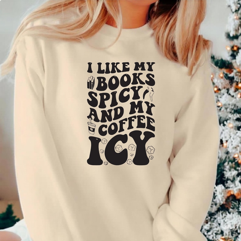 May include: Cream-colored sweatshirt with the text "I LIKE MY BOOKS SPICY AND MY COFFEE ICY" in a black, bold font. The design includes book, chili pepper, coffee cup, and ice cube graphics. A casual, comfortable top.