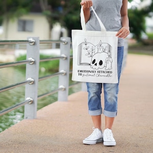 May include: A white tote bag with a black graphic of a skull, candles, and an open book. The text "Emotionally Attached to Fictional Characters" is printed below the graphic. The bag has two white handles.