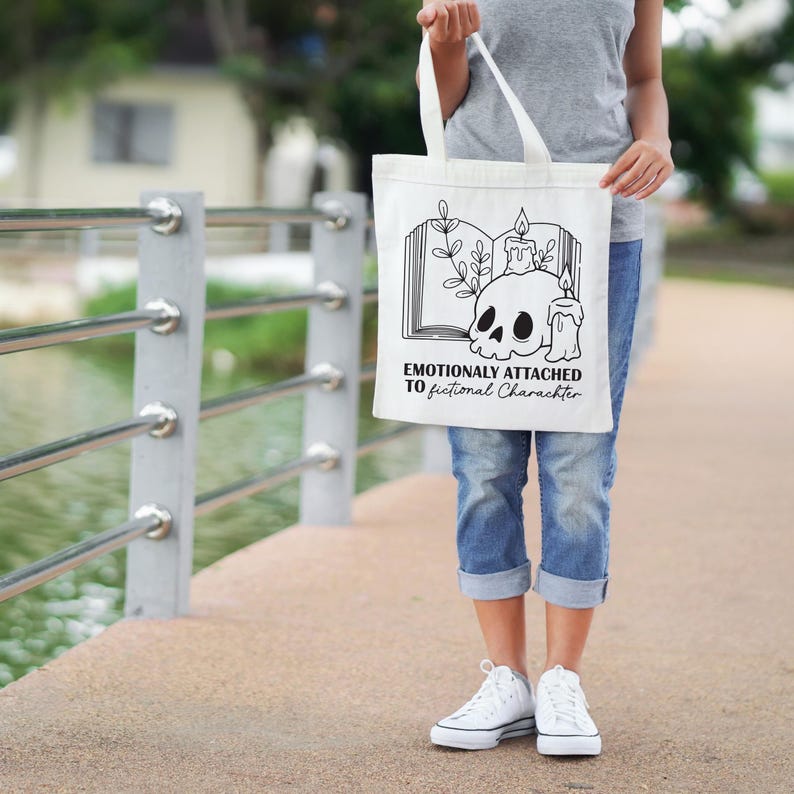 May include: A white tote bag with a black graphic of a skull, candles, and an open book. The text "Emotionally Attached to Fictional Characters" is printed below the graphic. The bag has two white handles.