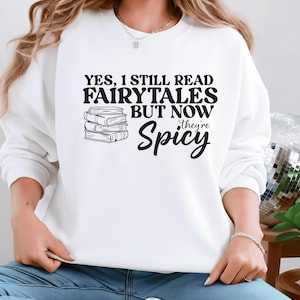 May include: White sweatshirt with the text "YES, I STILL READ FAIRYTALES BUT NOW they're Spicy" in black. A stack of books is to the left of the text. The sweatshirt is a long-sleeved crew neck.