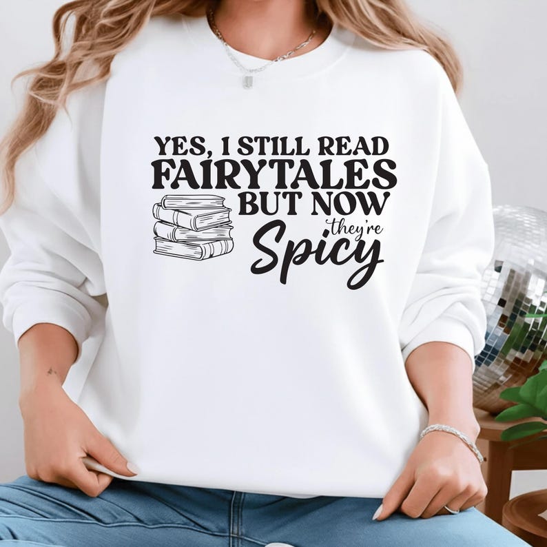 May include: White sweatshirt with the text "YES, I STILL READ FAIRYTALES BUT NOW they're Spicy" in black. A stack of books is to the left of the text. The sweatshirt is a long-sleeved crew neck.