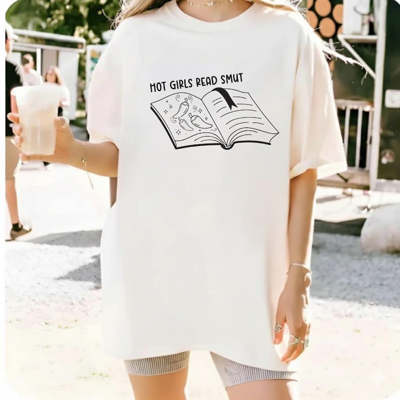 May include: Cream-colored t-shirt with the text "HOT GIRLS READ SMUT" above a black illustration of an open book with chili peppers and sparkles. The shirt is a casual, oversized fit.