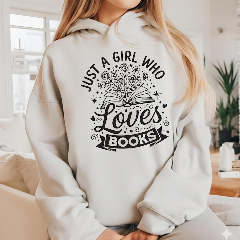 May include: A light beige hoodie with the text "JUST A GIRL WHO LOVES BOOKS" in black. The design includes an open book with flowers and stars. The hoodie has a drawstring hood and long sleeves.