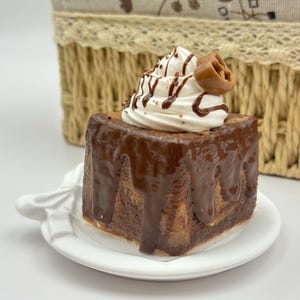 May include: A square chocolate cake with chocolate frosting and whipped cream topping, drizzled with chocolate sauce. The cake sits on a white plate with a decorative handle. A woven basket is in the background.