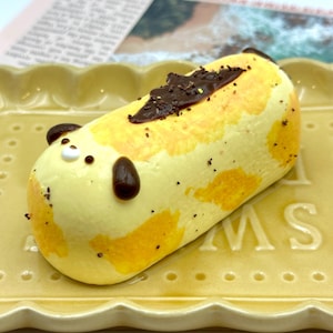 May include: A yellow pastry shaped like a bear, with brown ears and chocolate details, sits on a yellow rectangular plate. The pastry has yellow and orange spots and is sprinkled with dark chocolate flakes.