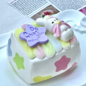 May include: A white, cake-shaped decorative item with pastel star and swirl accents. A purple star with the text "Do what you want" sits atop the cake, alongside a white bunny figure wearing a pink scarf. The cake is on a white plate.