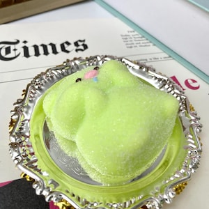 May include: A lime green marshmallow treat with a pink and blue accent on a silver and gold decorative plate. The plate is on a newspaper with the word "Times" visible.