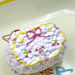 May include: A decorative, cloud-shaped item featuring a cartoon bunny face, a pink bow, and yellow and pink detailing. The item is adorned with small hearts and cherries. It rests on a light yellow surface.