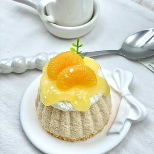 May include: A small dessert on a white plate, topped with orange slices and yellow glaze. The dessert sits on a textured, beige base with white cream. A small white creamer and spoon are in the background.