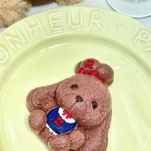 May include: A brown teddy bear-shaped confection with a red bow, holding a blue, white, and red emblem. The bear is on a yellow plate with the word "BONHEUR" embossed. A blue and white striped object is in the background.