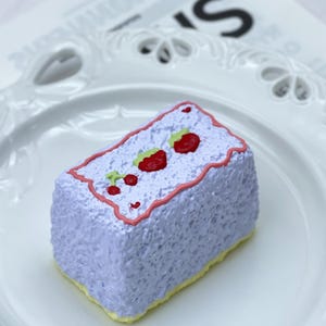 May include: A lavender-coloured rectangular cake with a textured surface. It is decorated with red strawberries and a pink border. The base of the cake is yellow. The cake sits on a white plate.