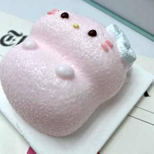 May include: A pink, textured cat figurine with brown eyes, a yellow nose, and pink cheeks. The cat has a white bow and is on a white surface. A collectible item.