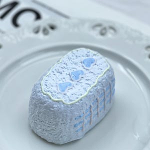 May include: A light blue, rectangular dessert with a textured surface, decorated with three light blue heart shapes on top. The sides feature vertical blue lines. The dessert sits on a white, scalloped-edge plate.
