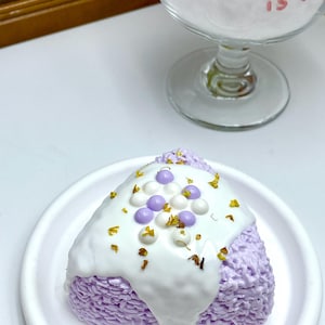 May include: A lavender-colored, triangular-shaped decorative item, possibly a candle or soap, on a white plate. It is topped with white frosting, purple and white spherical decorations, and small yellow accents. A glass with white fluffy material is in the background.