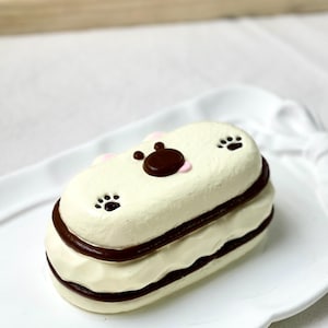 May include: A cream-coloured, bear-shaped dessert with chocolate accents and paw prints. The dessert is on a white plate with a scalloped edge. The bear's face has a dark brown nose and eyes, and pink ears.