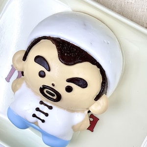 May include: A cartoon chef figurine with a white hat and uniform, paired with blue trousers. The figurine has a tan face, black eyes and hair. It is placed on a white tray with a red accent.