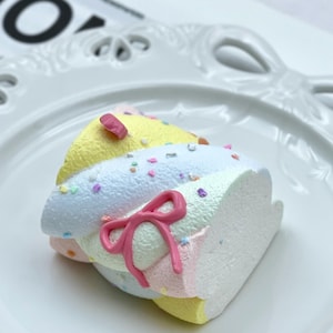 May include: A multi-layered dessert in pastel shades of yellow, blue, pink, and white, adorned with sprinkles and a pink bow. The dessert sits on a white, decorative plate.