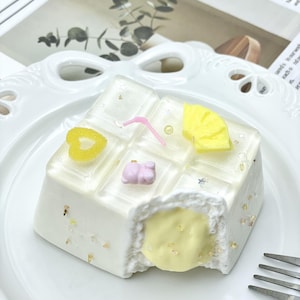 May include: A square white dessert with a bite taken out, revealing a yellow filling. The top is decorated with yellow and pink accents, and gold flakes. The dessert sits on a white plate with a fork.