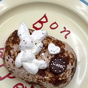 May include: A decorative dessert on a plate. The oval-shaped dessert has a brown and tan exterior, topped with a white bunny figure, a white heart, and a chocolate disc with the word "Catti". The plate has red text and a blue stripe.